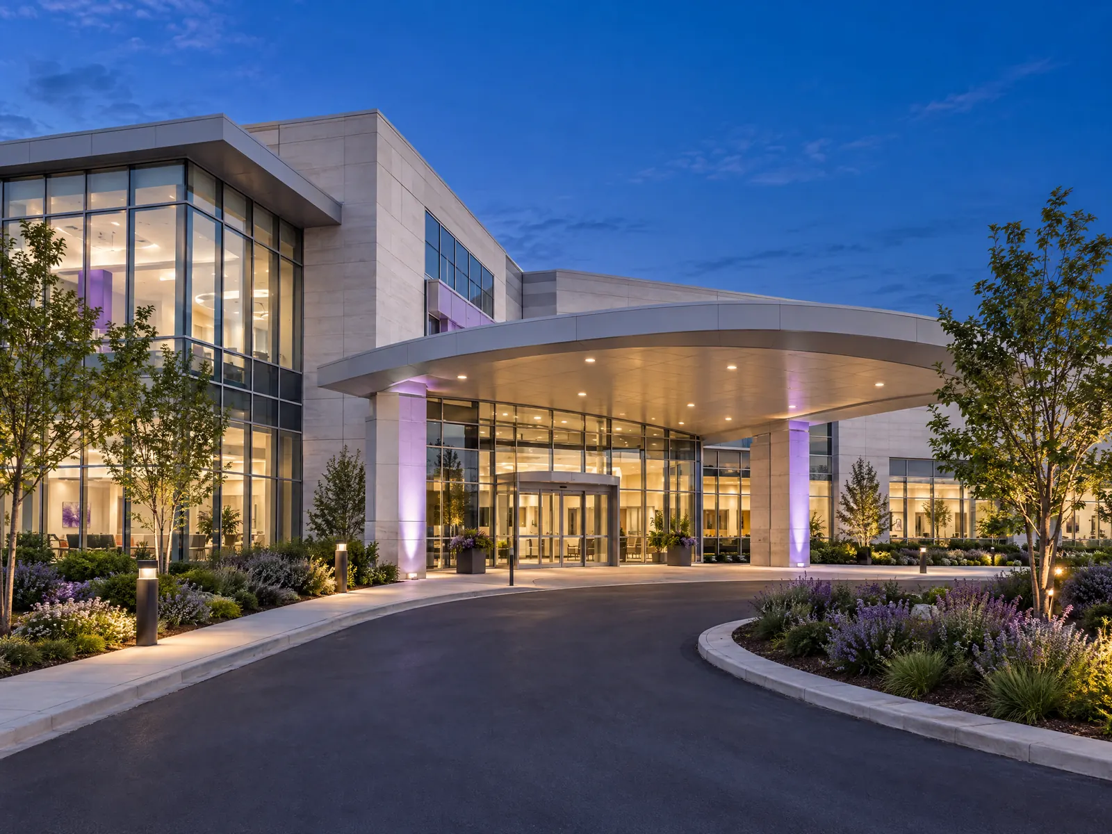 Glass hospital pavilion with a curved entry canopy and drop off loop at dusk